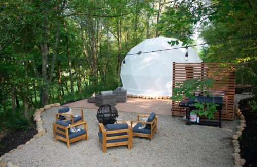 A cozy outdoor seating area with wooden chairs and a fire pit, next to a geodesic dome in a wooded setting.