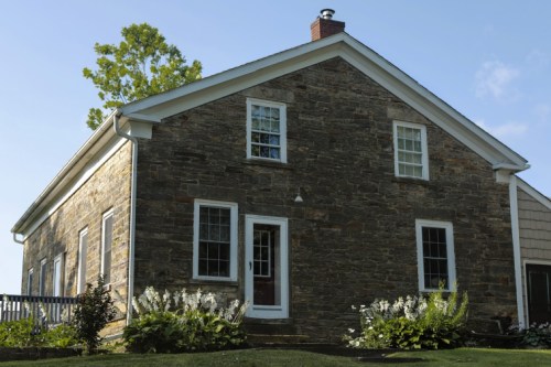 A stone house with white trim, surrounded by greenery and flowers, under a clear blue sky.