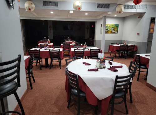 A spacious restaurant interior with round tables set for dining, featuring red tablecloths and black chairs.