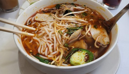 A bowl of spicy noodle soup with vegetables, herbs, and seafood, served with chopsticks.