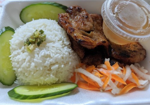 A takeout container with grilled meat, rice, cucumber slices, and pickled vegetables, accompanied by a sauce.