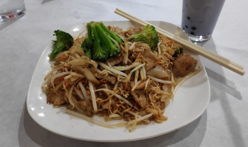 A plate of stir-fried noodles with vegetables and chicken, accompanied by a drink with tapioca pearls.