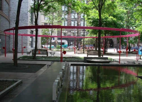 A circular pink structure in a park with benches, trees, and a reflective pond in the foreground.