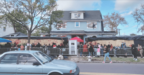 A crowd gathers outside a boarded-up house with Halloween decorations, while a car is parked nearby on the street.