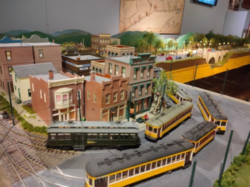 A detailed model train setup featuring buildings, tracks, and vintage-style trams in a scenic landscape.