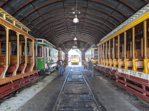 Two vintage streetcars inside a covered shed, with people walking between them on the tracks. Bright light at the end.