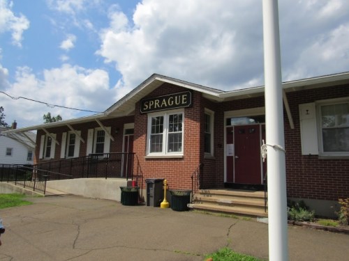 A brick building with a sign reading "SPRAGUE," featuring a ramp and steps, under a partly cloudy sky.