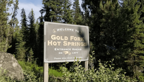 Sign welcoming visitors to Gold Fork Hot Springs, with directions to the entrance on the left. Surrounded by trees.
