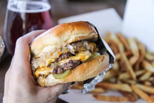 A hand holding a cheeseburger with onions and pickles, alongside a side of fries and a drink in the background.