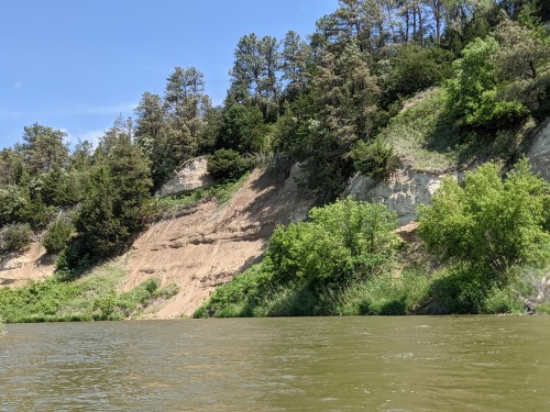 A serene river scene with lush green trees and a steep, sandy bank under a clear blue sky.