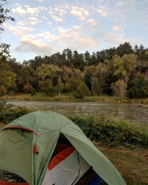 A green tent set up near a calm river, surrounded by trees and a cloudy sky.