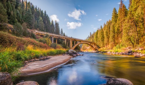 A serene river flows under a bridge, surrounded by lush green trees and a clear blue sky.