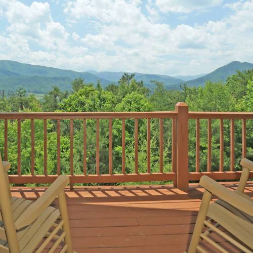A wooden deck with two chairs overlooks a lush green landscape and distant mountains under a partly cloudy sky.