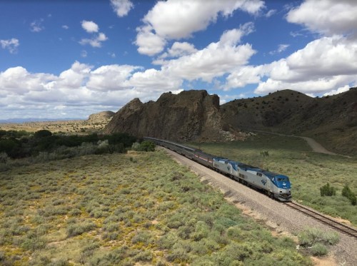 A train travels along a winding track through a green landscape with rocky hills and a blue sky dotted with clouds.