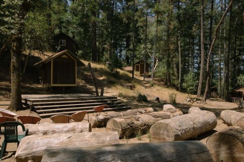 A serene forest scene with wooden cabins, logs, and a small stage surrounded by trees.