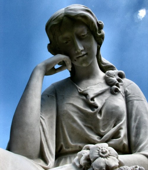 A serene marble statue of a woman resting her chin on her hand, surrounded by flowers against a blue sky.