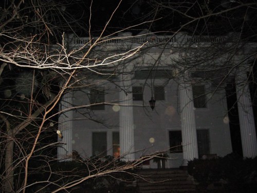 A dark, eerie house with white columns, partially obscured by bare branches and illuminated by a faint light.