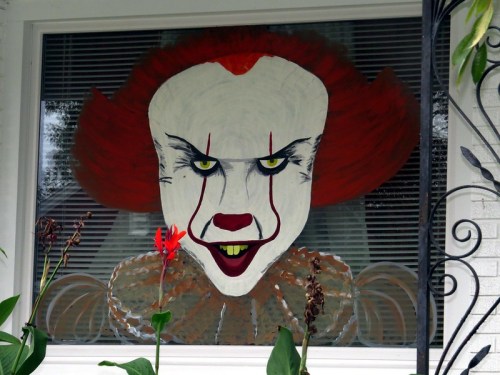 A large, painted depiction of a clown with red hair and menacing eyes in a window, surrounded by plants.