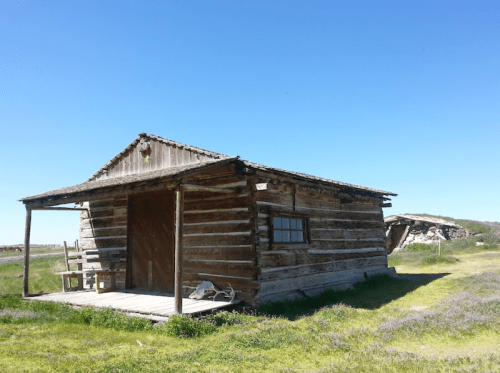 A rustic log cabin with a wooden porch, set against a clear blue sky and grassy landscape.