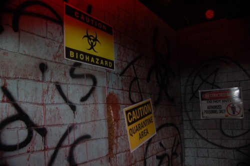 A dimly lit room with graffiti and caution signs warning of biohazards and quarantine areas.