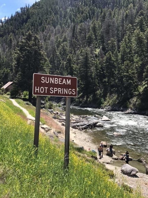 A sign for Sunbeam Hot Springs near a river, surrounded by lush green trees and a grassy area.