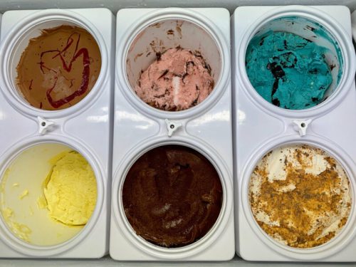 Six tubs of colorful ice cream: chocolate, strawberry, blue, yellow, brown, and a spiced flavor, arranged in a row.