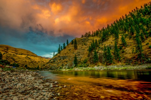 A serene river flows through a rocky landscape, surrounded by trees and illuminated by a colorful sunset sky.