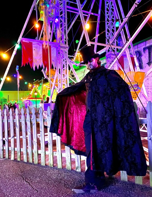 A man in a top hat and cape stands near a colorful carnival at night, with lights and a ferris wheel in the background.