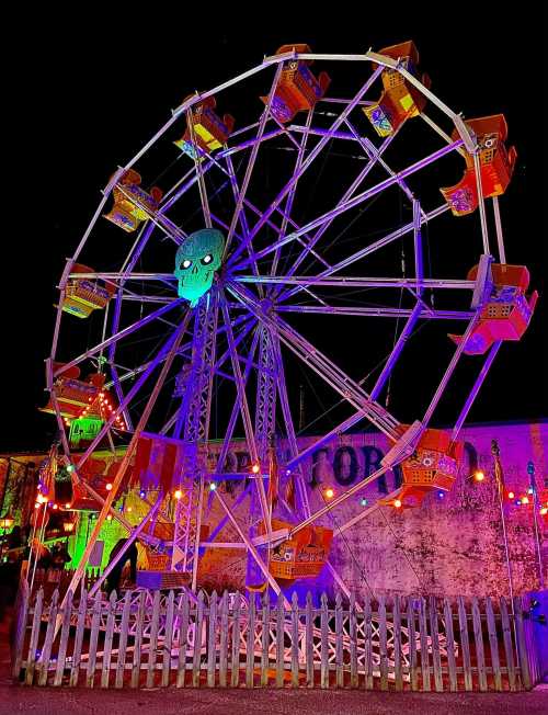 A colorful ferris wheel at night, featuring a skull at the top and illuminated by vibrant lights.