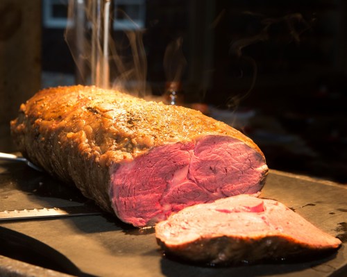 A perfectly cooked roast beef with a juicy pink center, steam rising, and a slice cut off to showcase its tenderness.