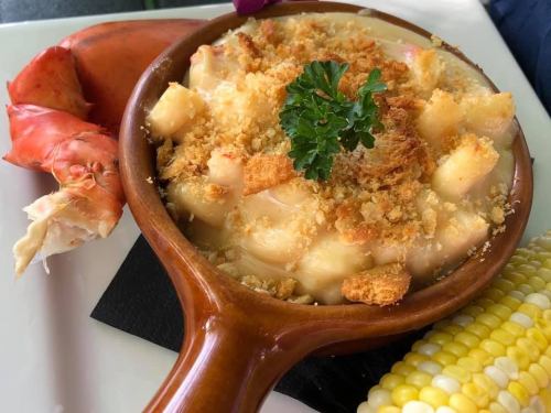 A creamy lobster mac and cheese topped with breadcrumbs, served with a lobster claw and corn on the side.