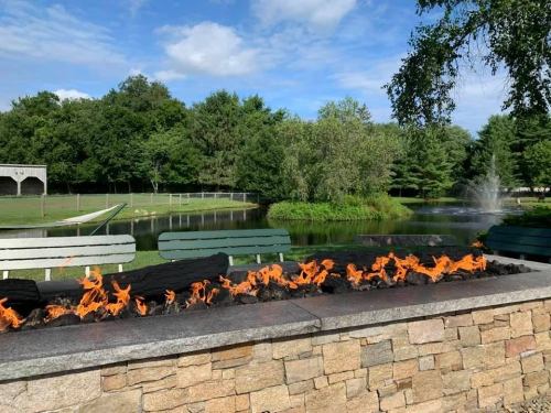 A stone wall with flames on top, overlooking a serene pond and lush greenery under a blue sky.
