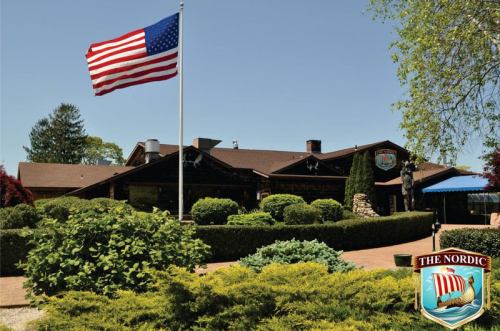 A rustic building with a flagpole displaying the American flag, surrounded by lush greenery and trees.