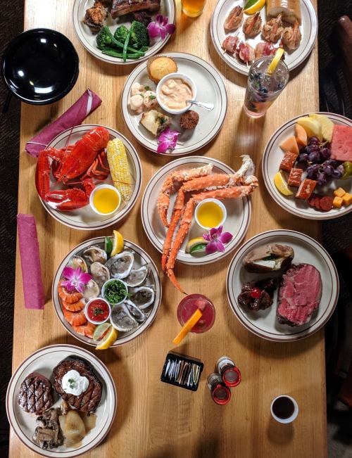 A variety of seafood and meats on plates, including lobster, crab, steak, and sides, arranged on a wooden table.