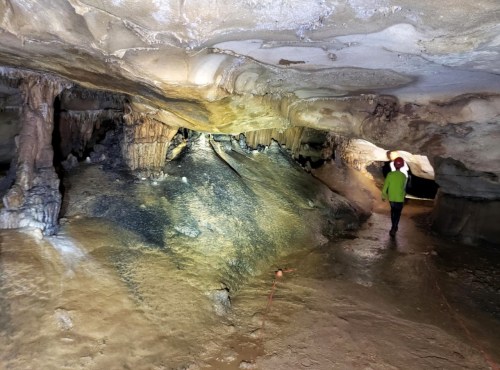 A person in a green shirt walks through a dimly lit cave with stalactites and stalagmites.