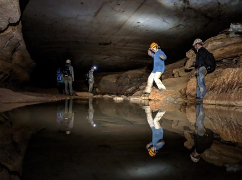Cavers explore a dark cave with headlamps, reflecting in a calm pool of water.
