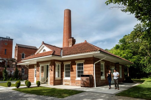 A brick building with a tall chimney, surrounded by greenery, with two people walking nearby.