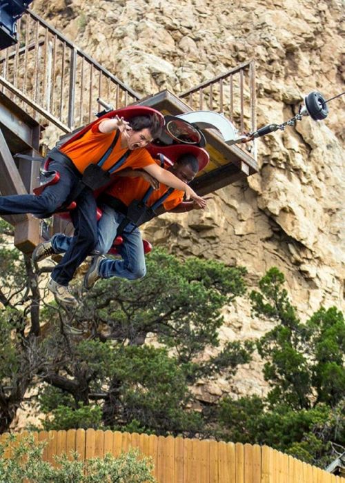 Two thrill-seekers in orange shirts are suspended in mid-air, about to drop from a ride against a rocky backdrop.