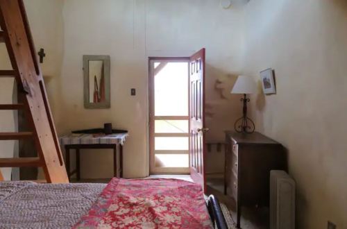 Cozy bedroom with a wooden ladder, a bed with a patterned blanket, a table, and a door leading to a bright hallway.