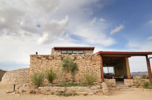 A modern stone house with a red roof, surrounded by desert landscape and sparse vegetation under a cloudy sky.