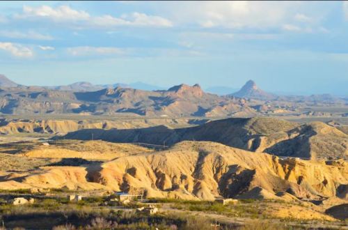 A panoramic view of rugged desert terrain with distant mountains under a blue sky and scattered clouds.