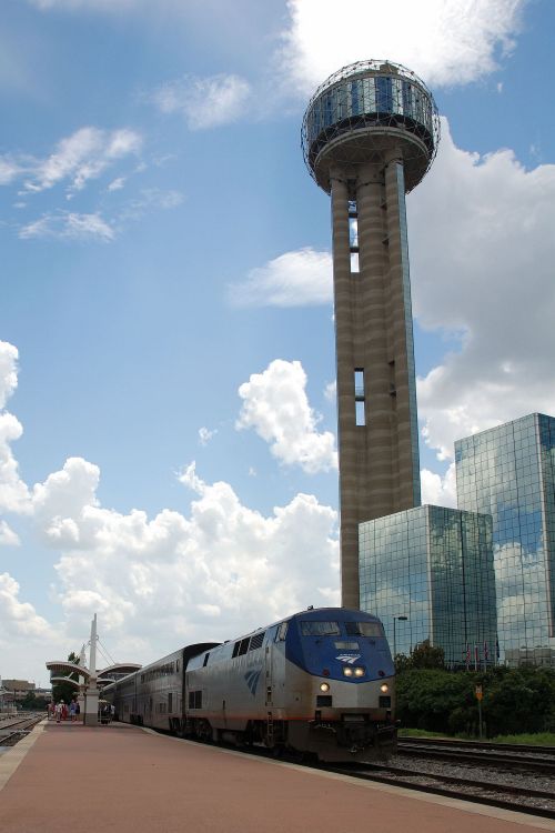 Amtrak train at a station with a tall tower and modern buildings under a partly cloudy sky.