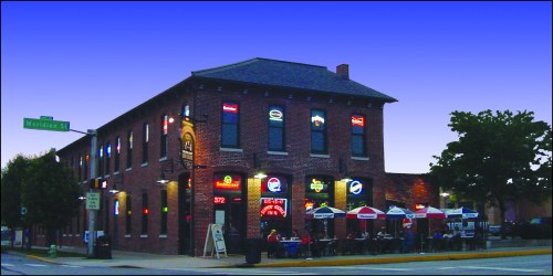 A brick building with colorful neon signs, featuring outdoor seating and umbrellas, at a street corner during twilight.