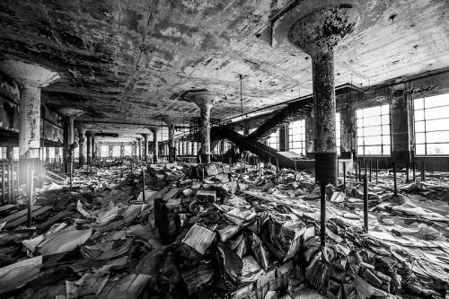 A dimly lit, abandoned warehouse filled with scattered papers and debris, featuring exposed pillars and large windows.