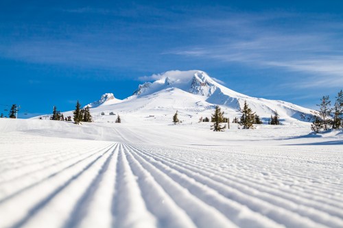 A snowy landscape with groomed ski tracks leading to a majestic mountain under a clear blue sky.