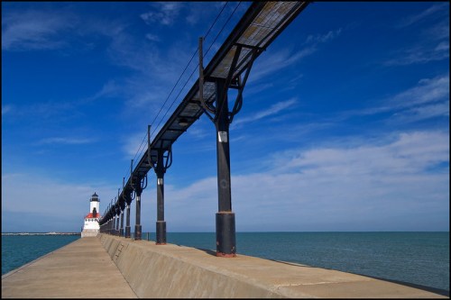 A lighthouse stands at the end of a long pier, with calm waters and a blue sky in the background.