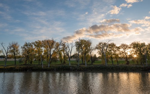 A serene river scene at sunset, with trees lining the bank and clouds in a colorful sky.