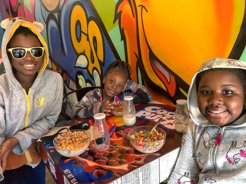 Three children smile at a table with colorful cereal bowls and milk, set against a vibrant graffiti background.