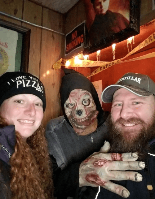A woman and a man pose with a person in a creepy mask and makeup, showcasing a spooky atmosphere.