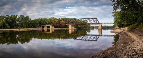 A tranquil river scene featuring a rusty train bridge reflecting in the water, surrounded by lush greenery.